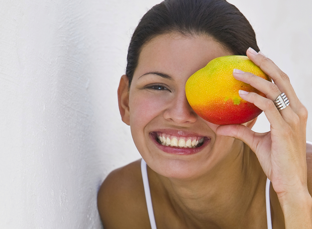 Woman holding mango in front of face outdoors, smiling, portrait