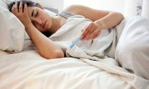 Eine Frau liegt im Bett, klammert sich an den Kopf und schaut auf ein Thermometer